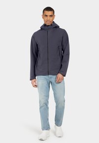 Grå vattentät jacka med framdragkedja, huva och två sidofickor; kombinerad med ljusblå jeans och vita sneakers.