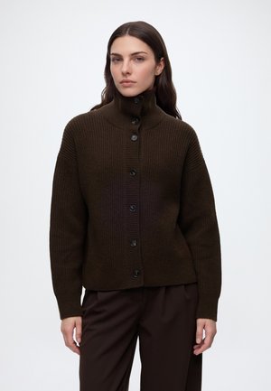 Frau mit langen braunen Haaren, die einen dunkelbraunen, gerippten Strickpullover mit Knöpfen und passende dunkle Hosen vor weißem Hintergrund trägt.