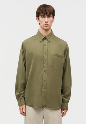 Νέος άνδρας που στέκεται, φοράει πουκάμισο σε ελαιόχρωμο με κουμπιά και τσέπη στο στήθος, καθώς και ανοιχτό μπεζ παντελόνι, μπροστά από απλό φόντο.