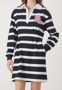 Randig klänning med marinblå och vita färger, polo-krage, långa ärmar och en rosa broderad "KL" logotyp på bröstet.