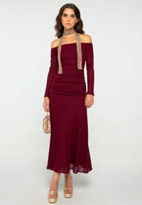 Robe ajustée bordeaux avec un design dénudé d'épaule, texture froncée et un nœud de cou marron clair. Associée à des talons compensés dorés et un sac beige.