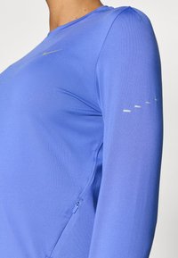 Μπλούζα Nike με μακριά μανίκια σε ζωηρό μπλε χρώμα, με απαλή υφή υφάσμ ατος, διακριτικές ραφές στους ώμους και αντανακλαστικές λεπτομέρειες στο μανίκι.