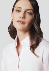 Jasnoróżowa koszula z guzikami z przodu, z usztywnionym kołnierzykiem, posiadająca dwa guziki przy szyi i gładką, teksturowaną tkaninę.