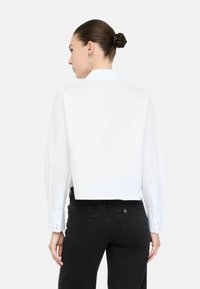 Camicia bianca cropped con maniche lunghe e polsini con bottone, presenta un colletto classico; abbinata a jeans neri, che mostrano una vestibilità slim.