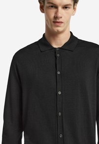 Svart knappupp sweater med polo-krage, långa ärmar och textilstruktur. Har sex knappar fram för stängning.