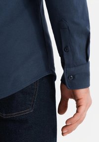 Marineblauwe katoenen langemouwen overhemd met knoopmanchetten en een rechte zoom. Gestructureerde stof met een gladde afwerking en zichtbare stiksels.
