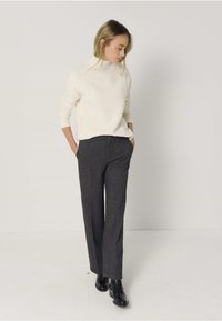 Kremowy dzianinowy sweter z golfem o fakturowanym wzorze, zestawiony z ciemnoszarymi spodniami o szerokich nogawkach. Model stoi z rękami w kieszeniach.