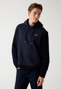 Felpa blu navy realizzata in tessuto morbido, con maniche a treccia, tasca a canguro e cappuccio con lacci dotati di terminali metallici.