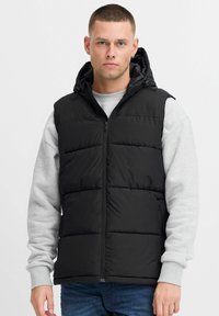 Schwarze Steppweste mit Kapuze, mit gesteppten Bereichen und einem Reißverschluss. Tragen über einem hellgrauen Sweatshirt und Jeans.