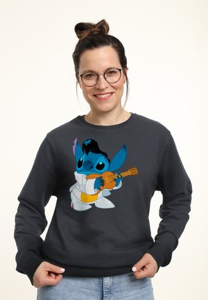 Kvinde med briller og håret sat op i en knold, iført en mørk sweatshirt med en tegneseriefigur, der spiller guitar, smilende, med hænderne om kanten af sweatshirten.