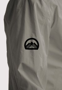 Giacca leggera bianca con un logo nero sulla manica sinistra, che presenta immagini di montagne con linee e forme geometriche.