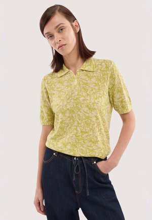 Kvinde iført en kortærmet grøn mønstret polo og mørkeblå jeans, stående med den ene hånd i lommen.