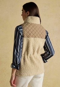 Kreemikas fliisvest koos neljakandilise pruuni ülaosaga, kombineeritud triibulise särgiga. Omab kõrget kaelust ja pehmet tekstuuri, sobib kihilise riietuse jaoks.