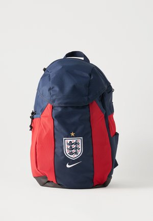 Navy plavi i crveni ruksak s grbom engleske nogometne reprezentacije, zlatnom zvijezdom i bijelim Nike logom sprijeda, stoji uspravno na jednobojnoj pozadini.