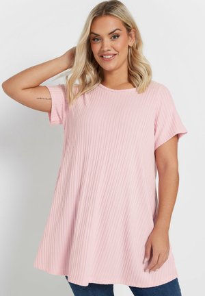 Uśmiechnięta kobieta o blond włosach, ubrana w luźny, prążkowany różowy top z krótkim rękawem i niebieskie dżinsy, pozująca z jedną ręką za głową.