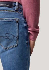 Hellblaue Jeans aus Denim mit strukturiertem Finish, ausgestattet mit einem Design für die Gesäßtasche und einem Markenzeichen. Kombiniert mit einem rosa Pullover.