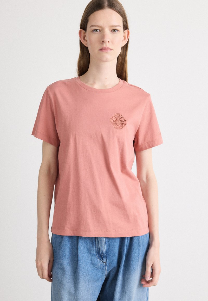 Tricou din bumbac roz, cu mâneci scurte și gât rotund, având un design embosat circular pe partea stângă a pieptului.