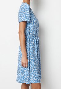 Žena v modrých šatech po kolena s bílými květinovými vzory, krátkými rukávy a páskem v oblasti pasu, stojící bokem na bílém pozadí.