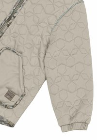 Manche et poche d'une veste matelassée beige clair avec couture géométrique en forme d'étoile, bordure florale contrastante, et détail de poignet élastique.