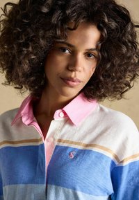 Kvinde med krøllet hår iført en hvid, blå og tan stribet skjorte med en pink krave, der kigger direkte ind i kameraet mod en neutral baggrund.
