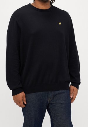 Homme portant un pull noir à manches longues avec un petit logo d'oiseau jaune sur la poitrine et un jean bleu foncé sur un fond blanc.