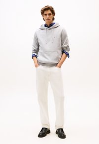 Γκρι βαμβακερό φούτερ με κουκούλα και μπροστινή τσέπη, συνδυασμένο με λευκό παντελόνι και μαύρα μοκασίνια. Το μοντέλο φοράει μπλε ριγέ πουκάμισο κάτω από το φούτερ.