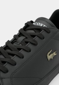 Sneaker in pelle nera con lacci piatti neri, cuciture del logo bianche e un accento dorato del logo. Texture liscia e design a profilo basso.