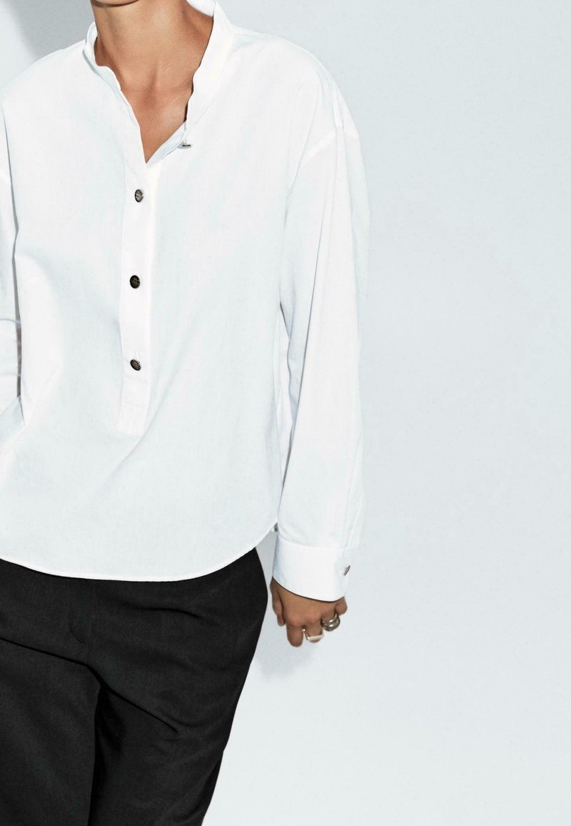 Personne portant une chemise blanche boutonnée avec manches retroussées et un pantalon noir, debout devant un fond gris clair.