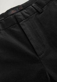 Czarne spodnie o gładkiej fakturze, z płaskim przodem, szlufkami na pasek i ukrytym zapięciem. Na pasku widać metki z etykietami.