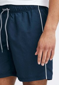 Costume da bagno blu navy con vita elastica, cordoncino e accenti laterali bianchi. Materiale liscio e leggero, che mette in mostra un design semplice.
