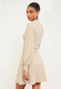 Beige stickad klänning med långa ärmar, texturerat diamantmönster, figursydd midja, utsvängd kjol och knappar på ärmsluten.