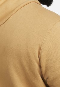 Bavlněná mikina v teplé béžové barvě, s jemnou strukturou a bočním pohledem na látku se švem na rameni.