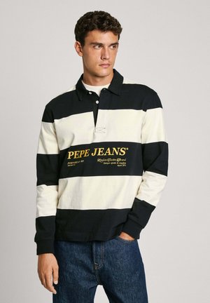 Schwarz-weiß gestreiftes Rugby-Shirt mit einem Kragen, ausgestattet mit einer Knopfleiste vorne und gelbem Logo-Text. Getragen mit dunkelblauen Jeans.