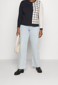 Woman wears black and white checkered cardigan, light blue flared jeans, black boots, and holds small white fuzzy handbag.