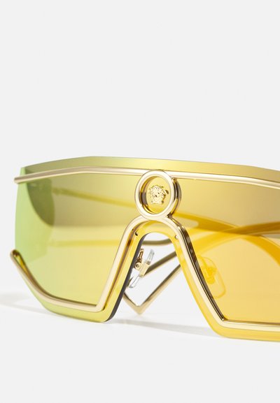 Occhiali da sole con montatura dorata e grandi lenti gialle sfumate. Presentano un design angolare audace e un prominente emblema di leone sulla montatura.