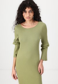 Ribbad, långärmad klänning i olivgrön. Har en rund ringning och en figurnära silhuett. Tyget verkar elastiskt och texturerat.
