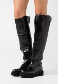 Stivali neri al ginocchio in pelle lucida con una robusta suola in gomma, dotati di una cerniera laterale e materiale tessurizzato.