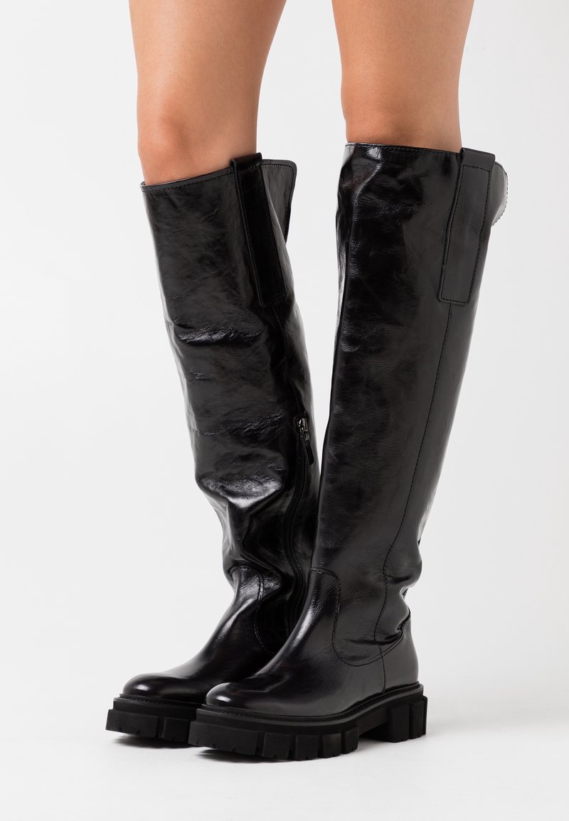 Musta polvipitkä kengät, jotka on valmistettu kiiltävästä nahasta paksulla kumipohjalla, sivuilla vetoketju ja teksturoitu materiaali.