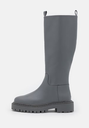 Botte haute en caoutchouc gris mat avec semelle épaisse à crampons et languettes à tirer en haut à l’avant et à l’arrière sur fond blanc.