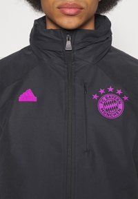 Personne portant une veste noire Adidas avec le logo violet du FC Bayern München et le logo Adidas sur la poitrine, fermeture éclair complètement fermée.