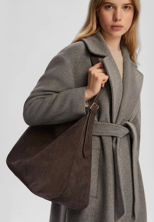 Borsa in suede marrone scuro con un design morbido e femminile, dotata di una lunga tracolla regolabile, che si abbina a un cappotto in lana grigia.