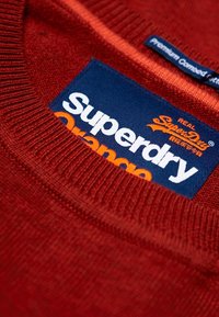Maglione rosso con colletto a coste. Presenta un'etichetta blu con "Superdry" in bianco e "Orange" in arancione, che indica il marchio e il nome del prodotto.