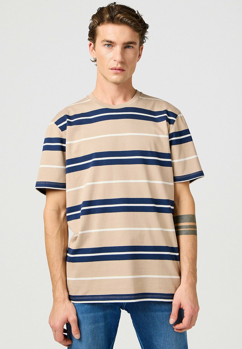 Camiseta a rayas beige con amplias rayas horizontales en azul marino y blanco, cuello redondo y mangas cortas, combinada con jeans azules.