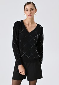 Maglione nero a V con motivo a diamante di paillettes argentate. Presenta maniche lunghe e orlo a coste. Abbinato a pantaloncini neri.