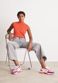 Orange ribbad ärmlös topp, ljusgrå vida jeans och flerfärgade sneakers med rosa och vita accenter, sittande på en kromstol.