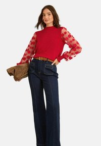 Piros kötött pulóver átlátszó virágmintás hímzett ujjakkal, sötétkék trapézfarmerrel és barna clutch táskával párosítva.