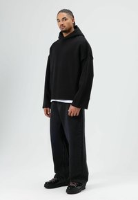 Zwarte oversized hoodie met capuchon, gedragen over een wit gelaagd shirt, gecombineerd met losse zwarte jeans en zwarte chunky sneakers met roze veters.