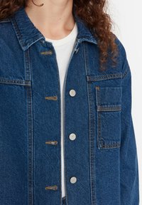 Tummansininen farkkutakki, jossa on napitettava etuosa, rintatasku ja kontrastivärinen ommel. Materiaali on teksturoitua ja takki on klassisessa muodossa.