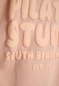 Ljusrosa sweatshirt med upphöjda, kraftiga bokstäver som står "PILATES STUDIO SOUTH BEACH EST. 1993." Mjuk, texturerad tyg.
