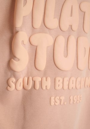 Lichtroze sweatshirt met verhoogde, stevige letters die "PILATES STUDIO SOUTH BEACH EST. 1993." zeggen. Zachte, textuurstof.